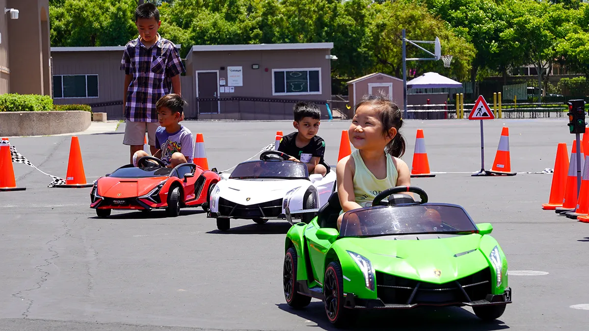 Three young kids smiling as they drive green, white and red Little Racers OC branded ride-on Lamborghinis around a coned racetrack with checkered flags and a traffic sign at an Orange County event