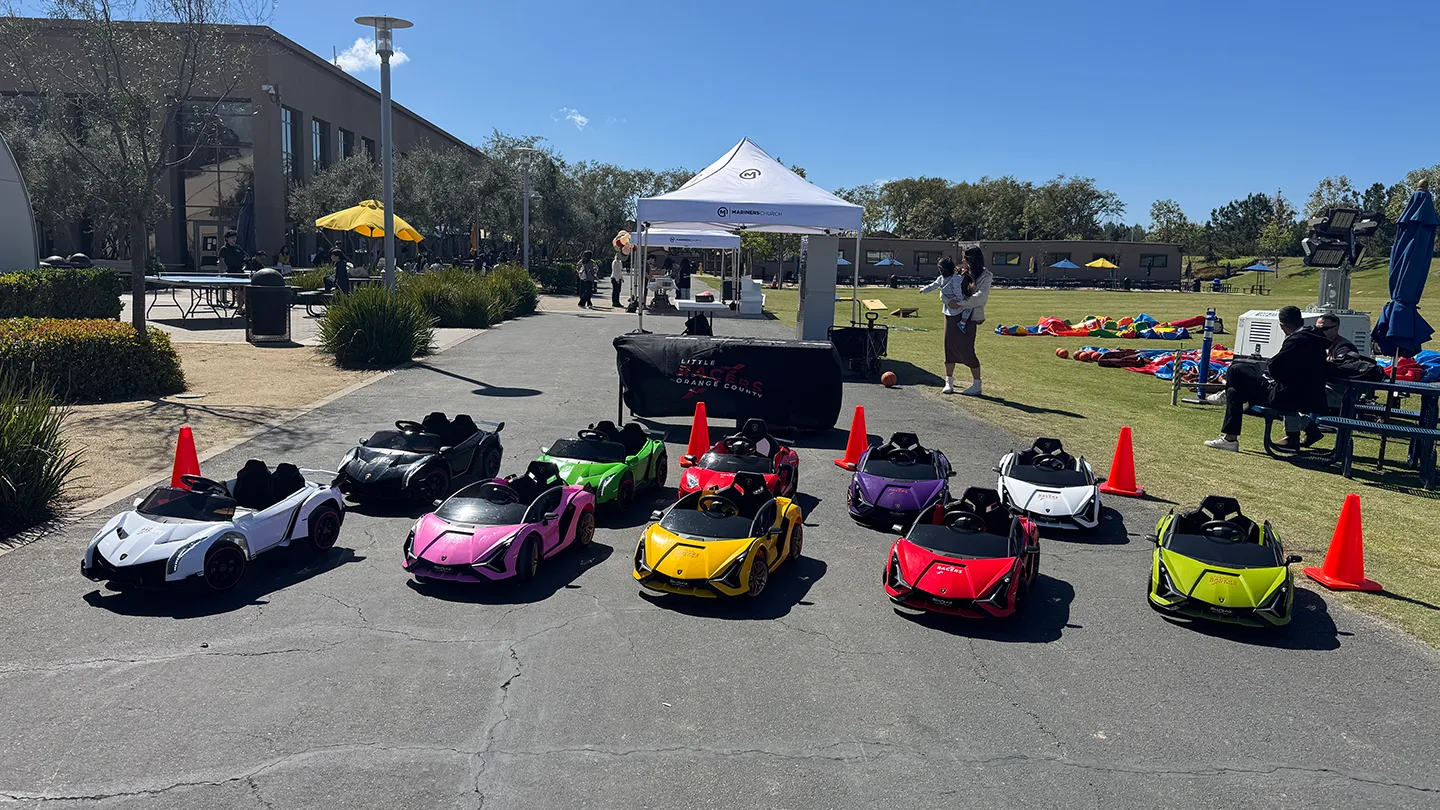 The full Little Racers OC fleet of branded kid-size ride-on Lamborghinis in white, black, green, pink, yellow, red, purple and lime lined up beside a Little Racers Orange County table at a community event