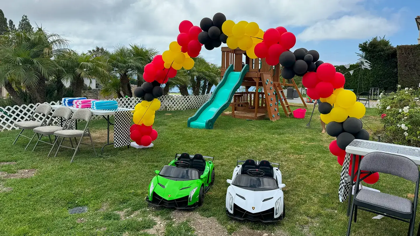 A backyard racing birthday party set up by Little Racers OC with a red, yellow and black balloon arch, checkered-flag table runners and green and white branded ride-on Lamborghinis