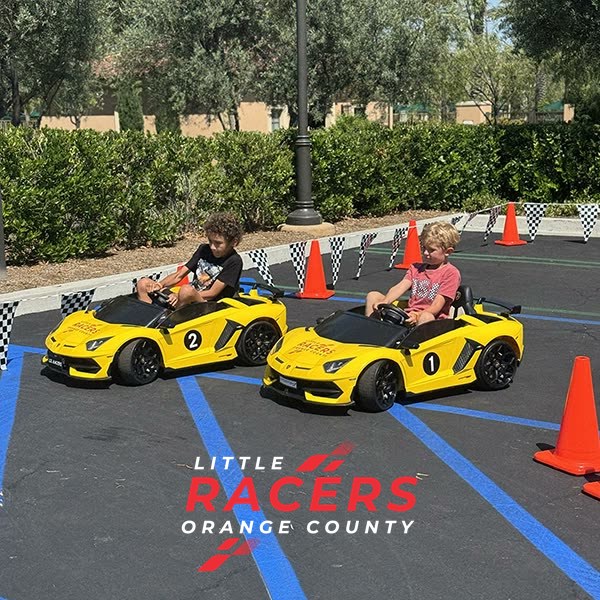 Two boys racing side by side in numbered yellow Little Racers OC branded ride-on Lamborghinis on a coned, checkered-flag racetrack
