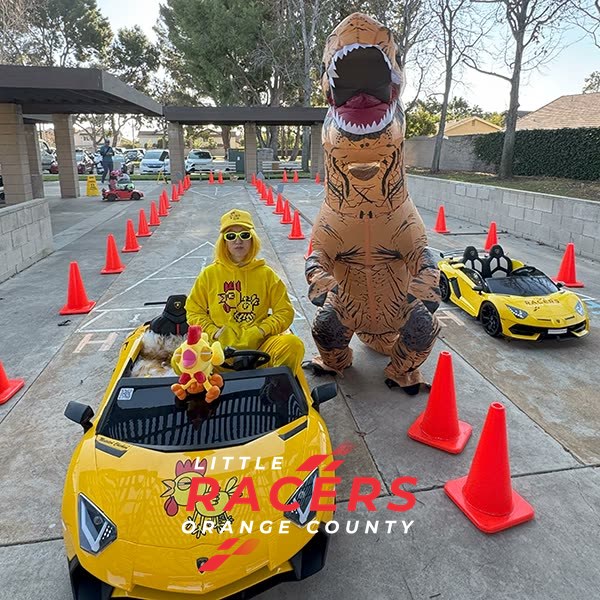 A child in a yellow costume driving a yellow ride-on Lamborghini next to a friend in an inflatable T-rex dinosaur costume on a coned track