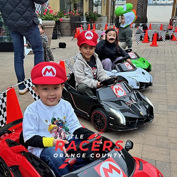 Three kids wearing Mario, Luigi and Peach costume hats racing red, black and white Little Racers OC branded ride-on Lamborghinis at a party