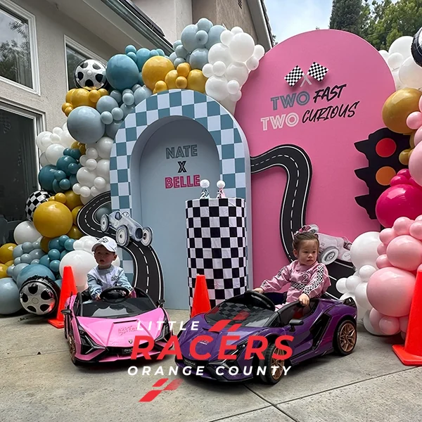 A 'Two Fast Two Curious' race-themed birthday party setup by Little Racers OC with a balloon arch and pink and purple branded ride-on Lamborghinis