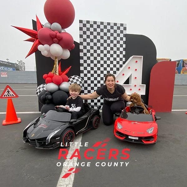 A parent and child crouched beside black and red branded ride-on Lamborghinis at a checkered number-four birthday backdrop